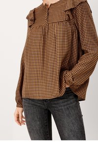 Blusa in gingham a scacchi marrone e nera con collo arruffato e maniche lunghe, realizzata in tessuto leggero. Tasche non visibili.