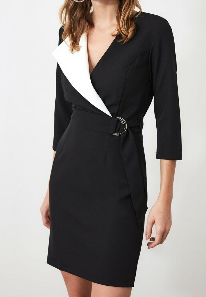 Femme portant une robe portefeuille noire avec un col à revers blanc et une ceinture à boucle double anneaux en argent, manches trois-quarts.