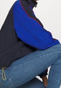 Person som knäböjer i en svart och kunglig blå jacka med röd piping och ljusblå denimjeans mot en vit bakgrund.