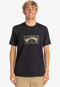 Black cotton t-shirt featuring a central logo patch with "Billabong" in yellow and white, crew neck, short sleeves, and relaxed fit.