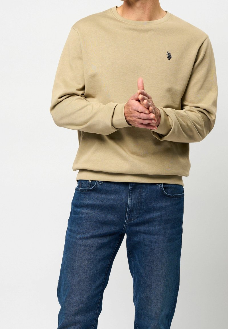 Homme portant un sweat-shirt beige avec un logo et un jean bleu, joignant les mains à la taille sur un fond clair uni.
