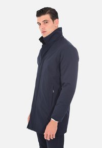 Impermeabile trench coat blu navy con colletto alto, dotato di zip frontale e tasche laterali, il tessuto ha una texture liscia e opaca.