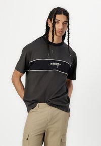 Dark gray t-shirt with a black horizontal stripe and white piping, featuring a white logo print. Paired with beige cargo pants.