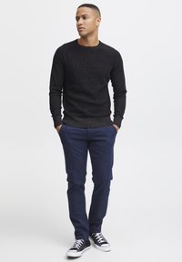 Zwarte gebreide trui met een textuurpatroon, gecombineerd met marineblauwe broek. Zwarte sneakers met witte zolen maken de outfit af.