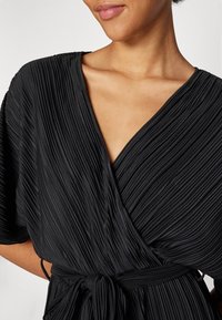 Robe noire plissée avec un décolleté en V et des manches courtes. Présente une ceinture assortie à nouer et un tissu lisse et texturé.