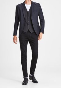 Musta bleiseri yhdistettynä vastaavaan liiviin ja valkoiseen T-paitaan, yhdessä mustien slim-fit-farkkujen ja mustien pukukenkien kanssa.