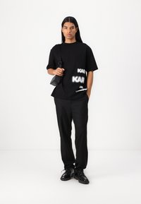 Μαύρο T-shirt με λευκά γραφικά σχέδια, συνδυασμένο με μαύρο παντελόνι. Το μοντέλο κρατάει μια μικρή μαύρη τσάντα και φοράει μαύρα sneakers, στέκεται σε λευκό φόντο.