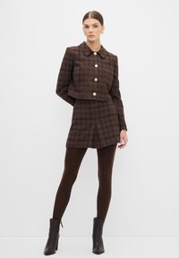 Giacca a quadrati marrone cropped con grandi bottoni abbinata a una gonna mini coordinata. Collant marroni e stivaletti con tacco completano l'outfit.