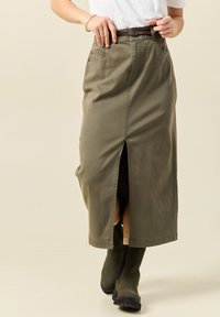 Khakifarbener Midirock mit einem Schlitz vorne, aus Baumwollstoff gefertigt, mit Gürtelschlaufen und Seitentaschen, kombiniert mit kniehohen Stiefeln.