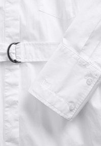 Jachetă albă impermeabilă, cu talie prevăzută cu curea, un buzunar la piept și manșete cu nasturi, având detaliu de logo brodat pe mânecă.