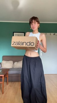 Hnedá kartónová krabica s potlačou "zalando" v čiernej farbe, držená pred osobou v bielej bezrukávovej blúzke a vo voľných čiernych nohaviciach.