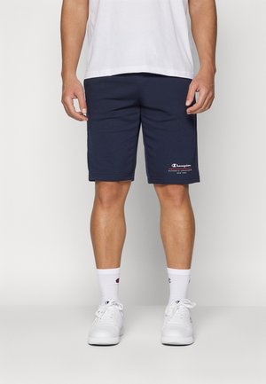 Pantalones cortos de algodón azul marino con bolsillos laterales, cinturilla elástica y un pequeño logo blanco de Champion en la pierna izquierda. Se llevan con zapatillas blancas y calcetines.