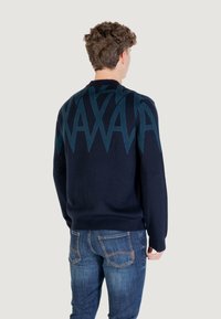 Un homme portant un pull bleu foncé avec un motif géométrique teal sur le haut du dos et un jean bleu, se tenant de dos sur un fond clair.