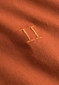 Oransje stoff med fin tekstur som har et brodert "II" i en lysere oransje tråd. Et nærbilde viser den detaljerte sømmen.