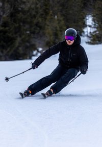 Skileraar in een zwarte jas en broek, met een zwarte helm en roze goggles, navigeert door de sneeuw met skistokken, op een besneeuwde helling.