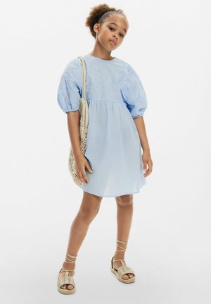 Robe bleu clair avec des manches bouffantes, un corsage brodé et une jupe fluide. Associée à un sac tissé et des sandales à lanières. Tissu lisse.