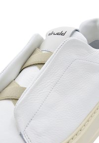 Biały skórzany sneaker z teksturowaną powierzchnią, kremowym elastycznym paskiem i wypukłym logo na pięcie. Zaokrąglony nosek i przeszycia jako detale.