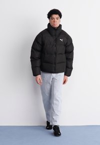 Chaqueta de plumas negra con cuello alto y cierre de cremallera, con un logo blanco de Puma en el lado izquierdo del pecho, combinada con unos jeans de color azul claro y zapatillas negras.