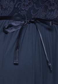 Marineklänning med spetsliv och plisserad kjol, samt ett satinförband i midjan. Tyget har en mjuk, texturerad yta.