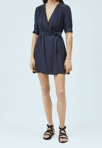 Navy modré zavinovací šaty s krátkými puff rukávy, výstřihem do V a zavázaným pasem. V kombinaci s černými sandály na tenkých páscích na nahých nohách.