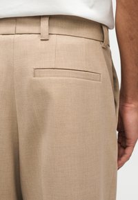 Μπεζ παντελόνι ραμμένο στα μέτρα με πίσω τσέπη με λάστιχο και θηλιές για ζώνη, φορεμένο με λευκό τοπ, εμφανίζεται από τη μέση έως το μέσο του μηρού.