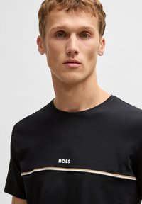 Schwarzes Baumwoll-T-Shirt mit Rundhalsausschnitt, kurzen Ärmeln und einem weißen "BOSS"-Logo über einem horizontalen Streifen in Gold und Weiß.