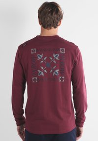 Chemise à manches longues bordeaux avec un dos à motifs en bleu et gris, comprenant des motifs floraux et le mot "OXBOW". Matière en coton doux.