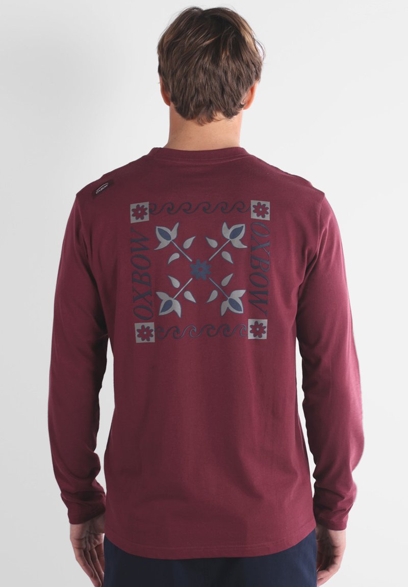 Chemise à manches longues bordeaux avec un dos à motifs en bleu et gris, comprenant des motifs floraux et le mot "OXBOW". Matière en coton doux.
