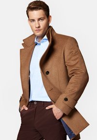 Brązowy wełniany płaszcz z wysokim kołnierzem, z podwójnym zapięciem na guziki i kieszeniami bocznymi, noszony na jasnoniebieskiej koszuli i ciemno-brązowych spodniach.