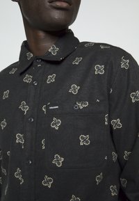 Homme portant une chemise noire à boutons avec des motifs paisley beiges et une poche poitrine boutonnée étiquetée "Volcom."