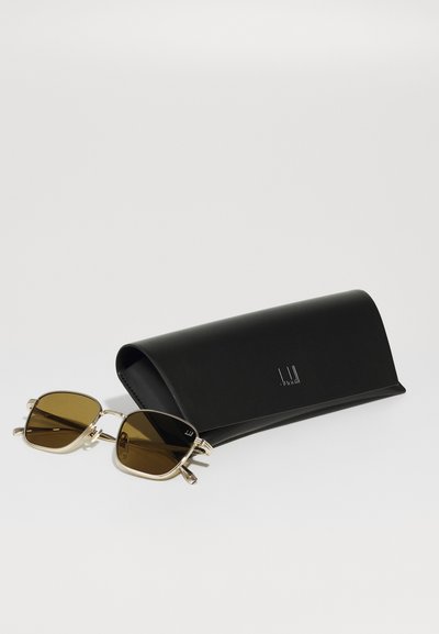Lunettes de soleil teintées brun avec une monture carrée et des accents dorés, posées à côté d'un étui en cuir noir portant un logo discret.