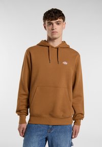 Giovane uomo che indossa un hoodie marrone con una tasca frontale e un logo, abbinato a jeans blu, in piedi davanti a uno sfondo grigio chiaro.