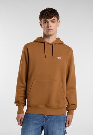 Giovane uomo che indossa un hoodie marrone con una tasca frontale e un logo, abbinato a jeans blu, in piedi davanti a uno sfondo grigio chiaro.