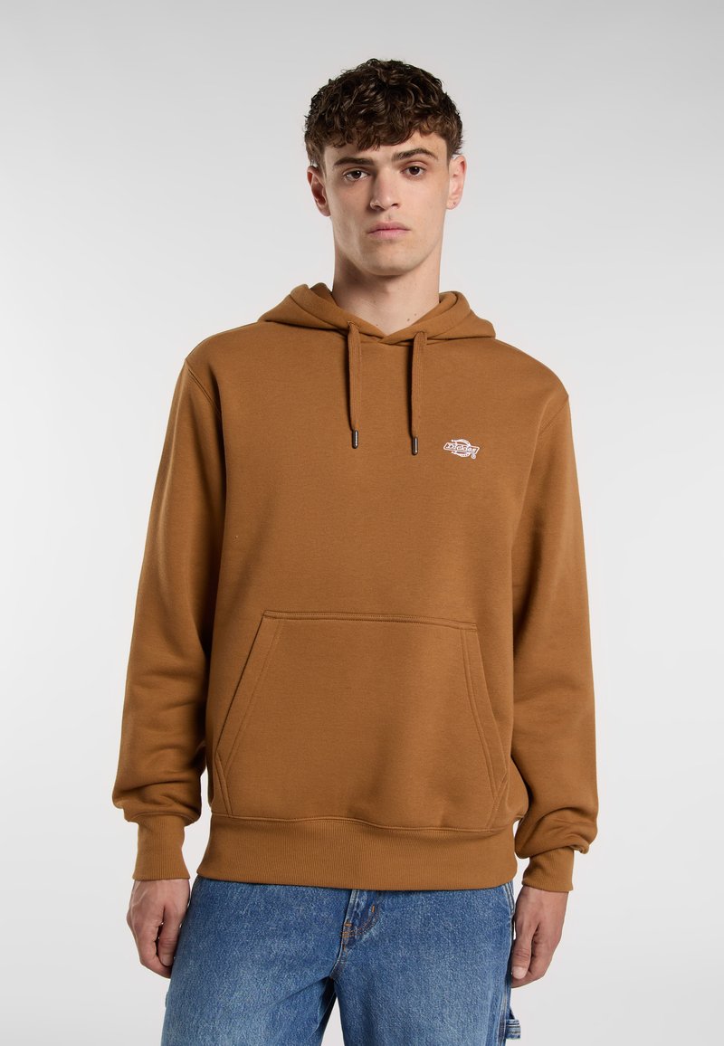 Giovane uomo che indossa un hoodie marrone con una tasca frontale e un logo, abbinato a jeans blu, in piedi davanti a uno sfondo grigio chiaro.