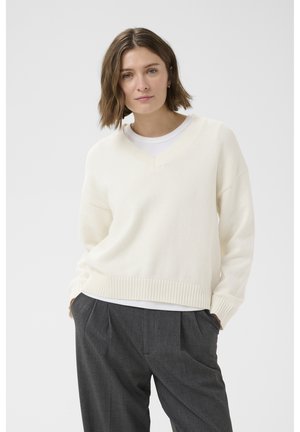 Žena s krátkymi hnedými vlasmi, oblečená v bielom svetri s výstrihom do V cez bielu košeľu a voľných sivých plisovaných nohaviciach, ruky v vreckách.