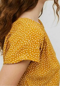 Blusa de color mostaza con lunares blancos, con mangas cortas y una textura sutil, realzada por un escote redondeado.