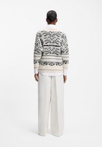 Γυναίκα που φαίνεται από πίσω, φορώντας πλεκτό πουλόβερ με ασπρόμαυρο μοτίβο, λευκό πουκάμισο με γιακά και λευκά παντελόνια με φαρδιά μπατζάκια, σε απλό φόντο.