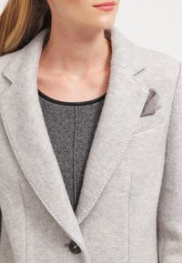 Jasnoszary filcowy blezer z klapami z wycięciem, zapinany na pojedynczy guzik, z dekoracyjną poszetką w kieszeni. Pod spodem ciemnoszary sweter.