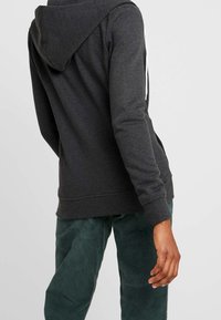 Muž v tmavě šedé mikině s kapucí a tmavě zelených manšestrových kalhotách, stojící bokem proti jednobarevnému světle šedému pozadí.