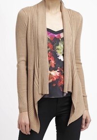Cardigan côtelé beige à design ouvert, manches longues et finition texturée ; porté sur un chemisier à motifs floraux et un pantalon noir.