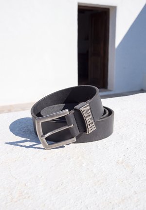 Un cinturón de cuero negro con una hebilla plateada y una trabilla con el logotipo de la marca reposa sobre una superficie de piedra blanca iluminada por el sol.