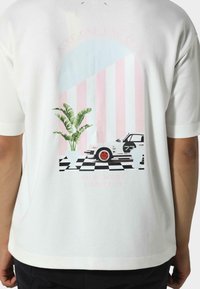 Valkoinen puuvillapaita, jossa on graafinen printti autosta, palmunlehvestä ja raidallisesta pastelitaustasta. Mukana mustavalkoisia ruutuelementtejä.