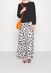 Schwarzes langärmliges Oberteil, lockere weiße Hose mit schwarzem Leopardenmuster und eine orangefarbene Umhänge Tasche. Riemchen schwarze Absatzschuhe runden den Look ab.