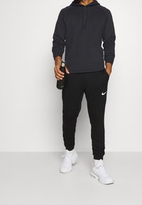 Felpa nera con cappuccio e dettagli grigi, abbinata a pantaloni della tuta neri con il logo Nike bianco. Completata con sneakers bianche.