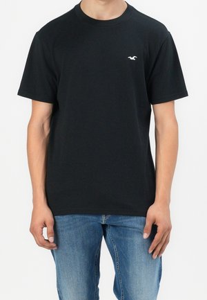 Zwarte katoenen t-shirt met korte mouwen en een ronde hals. Voorzien van een klein wit logo op de linkerborst. Draagt met blauwe denim spijkerbroeken.