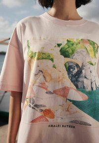Roza bombažna majica s pisano grafiko obale Amalfija z dežniki in skalnatimi pečinami, dopolnjena z napisom "AMALFI BATHER".