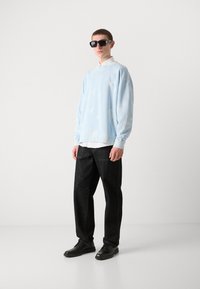 Sudadera de gran tamaño de color azul claro con un patrón de nubes, superpuesta a una camisa blanca con cuello, combinada con jeans de pierna ancha negros y zapatos negros.