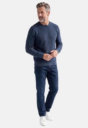 Mężczyzna z siwymi włosami i brodą, ubrany w granatowy sweter, ciemnoniebieskie dżinsy i białe trampki, stojący na tle jednolitego jasnego tła.