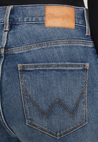 Modré džínsy s koženým nášivkom Wrangler, viditeľným šitím a výrazným dizajnom zadnej vrecka so vzorom v zigu-zagu.