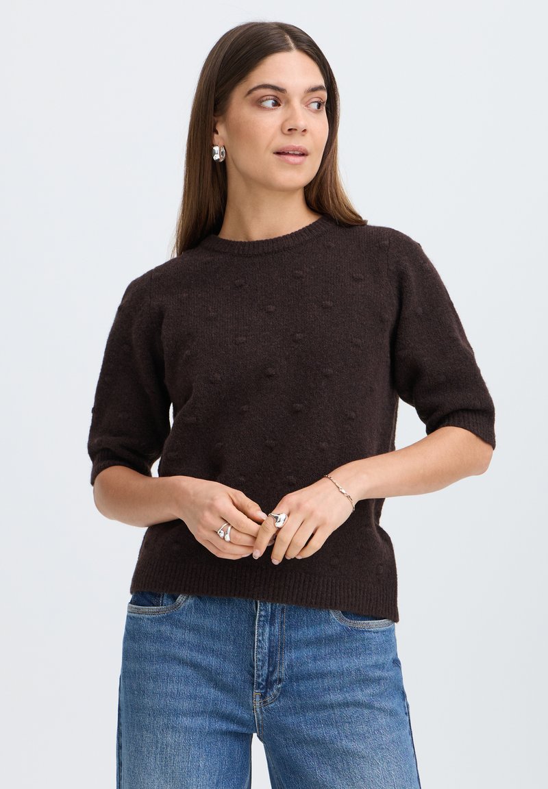 Femme aux longs cheveux bruns portant un pull texturé à manches courtes marron foncé et un jean bleu, regardant sur le côté les mains jointes.
